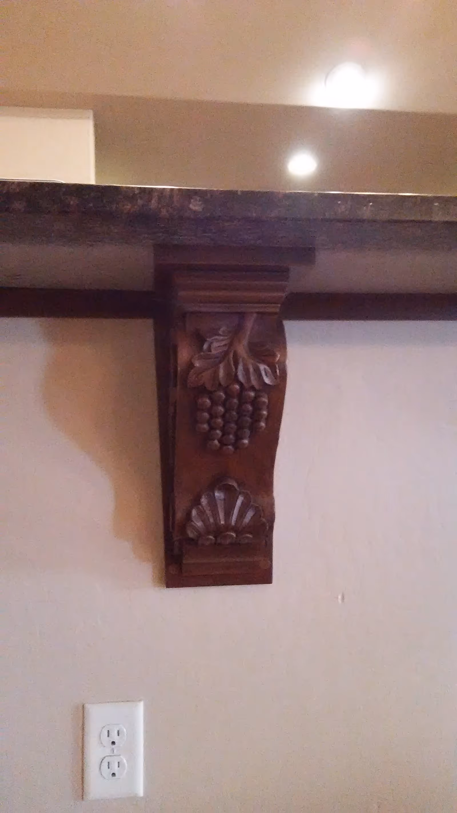 Close-up of a carved wooden corbel with grape and shell motifs supporting a countertop above a wall outlet.
