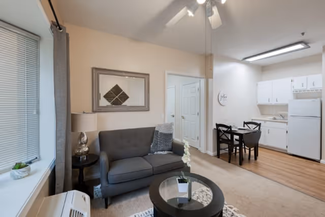 A cozy senior living apartment interior featuring a gray loveseat with a pillow, a round glass coffee table with a small plant, a side table with a lamp, and a large window with blinds and curtains. In the background, there is a small dining area with a black table and chairs, and a kitchenette with white cabinets, a refrigerator, and a sink. The walls are light-colored, and the floor is a combination of carpet and wood.