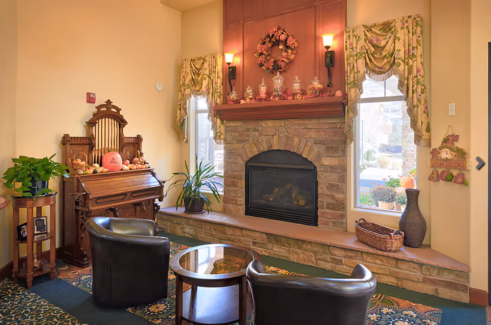 Cozy senior living room area with a stone fireplace decorated with autumn-themed items, two black leather chairs around a small round glass-top table, an antique wooden organ adorned with pumpkins, and windows with floral curtains letting in natural light.