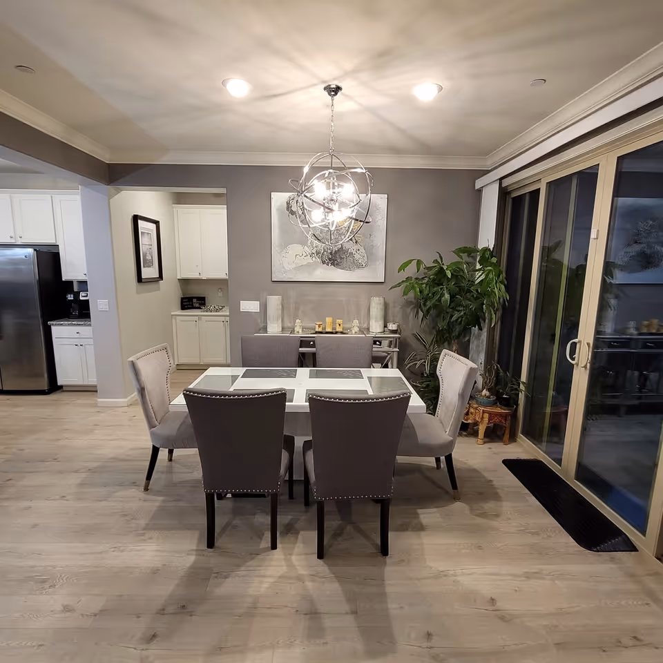 A modern dining area with a glass-top table and six upholstered chairs under a decorative chandelier, adjacent to sliding glass doors and an open kitchen.