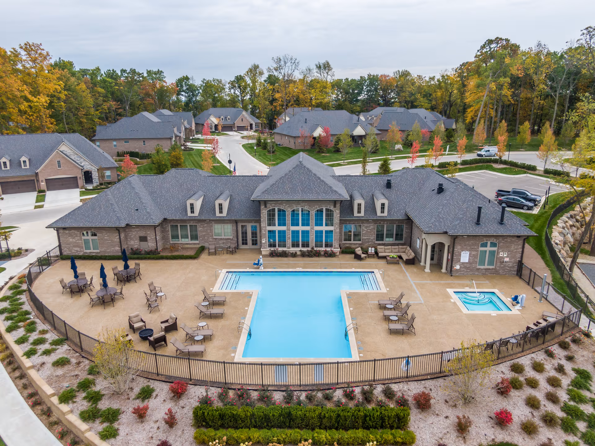Aerial view of a senior living facility featuring a large outdoor swimming pool and a smaller hot tub, surrounded by lounge chairs and tables with umbrellas. The building behind the pool has a brick exterior with large windows and a gray shingled roof. The area is fenced and landscaped with bushes and trees showing autumn colors. Several residential buildings and parked cars are visible in the background.