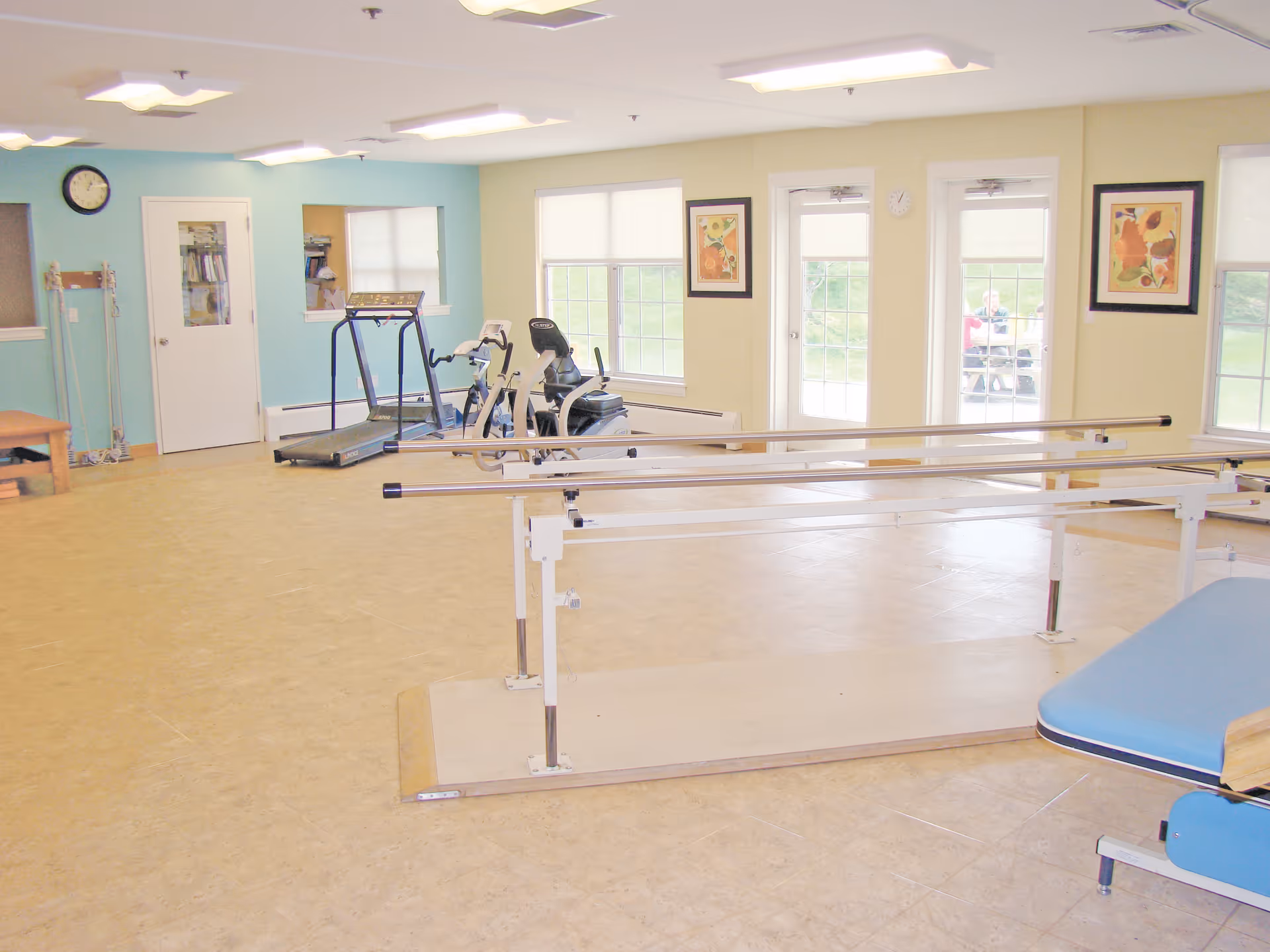 A spacious rehabilitation room with parallel bars for walking exercises in the foreground, exercise equipment including a treadmill and stationary bike against a light blue wall, large windows and glass doors letting in natural light, and framed artwork on the walls.