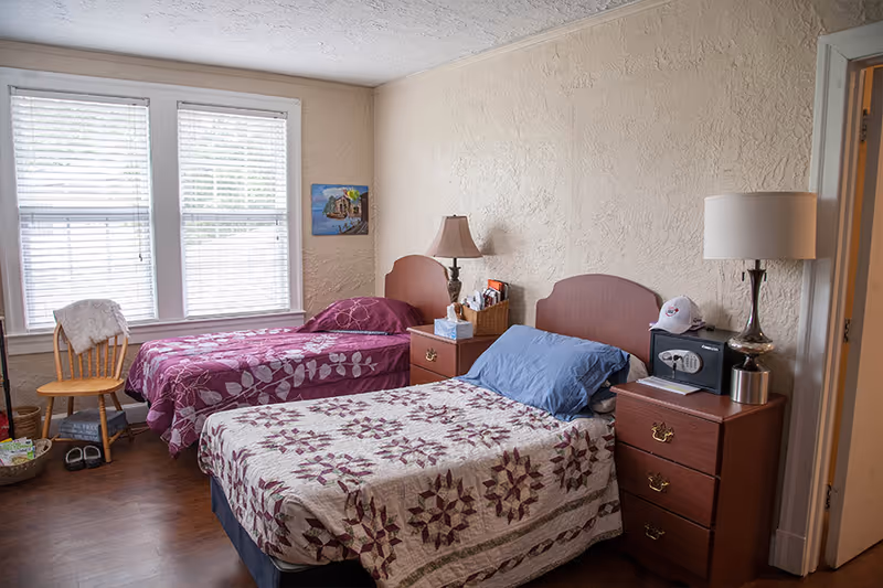 A cozy bedroom in an assisted living facility featuring two single beds with patterned bedspreads, a wooden chair near a window with blinds, two bedside tables each with a lamp, and a small safe on one of the tables.