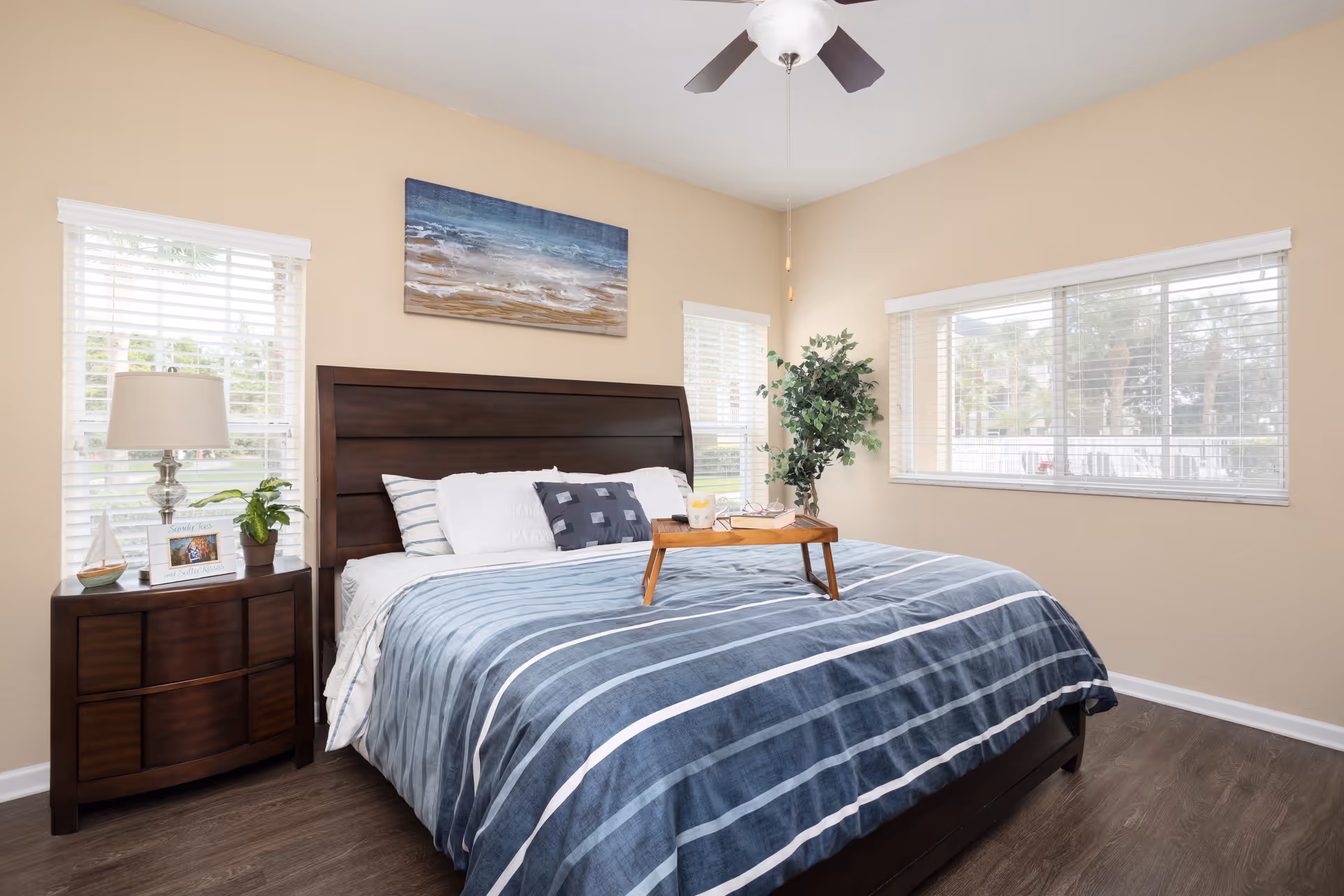 A bright bedroom with a large bed featuring a dark wooden headboard and blue striped bedding. There is a wooden tray on the bed with a drink and a book. The room has three windows with white blinds, a nightstand with a lamp, a small plant, and a framed photo. A ceiling fan with lights is above the bed, and a painting of a beach scene hangs on the wall.