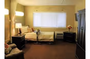 A cozy nursing home bedroom with a single bed against the wall, a bedside table with a lamp, a small table on wheels, a comfortable armchair, a window with closed blinds, and a television mounted on the wall.