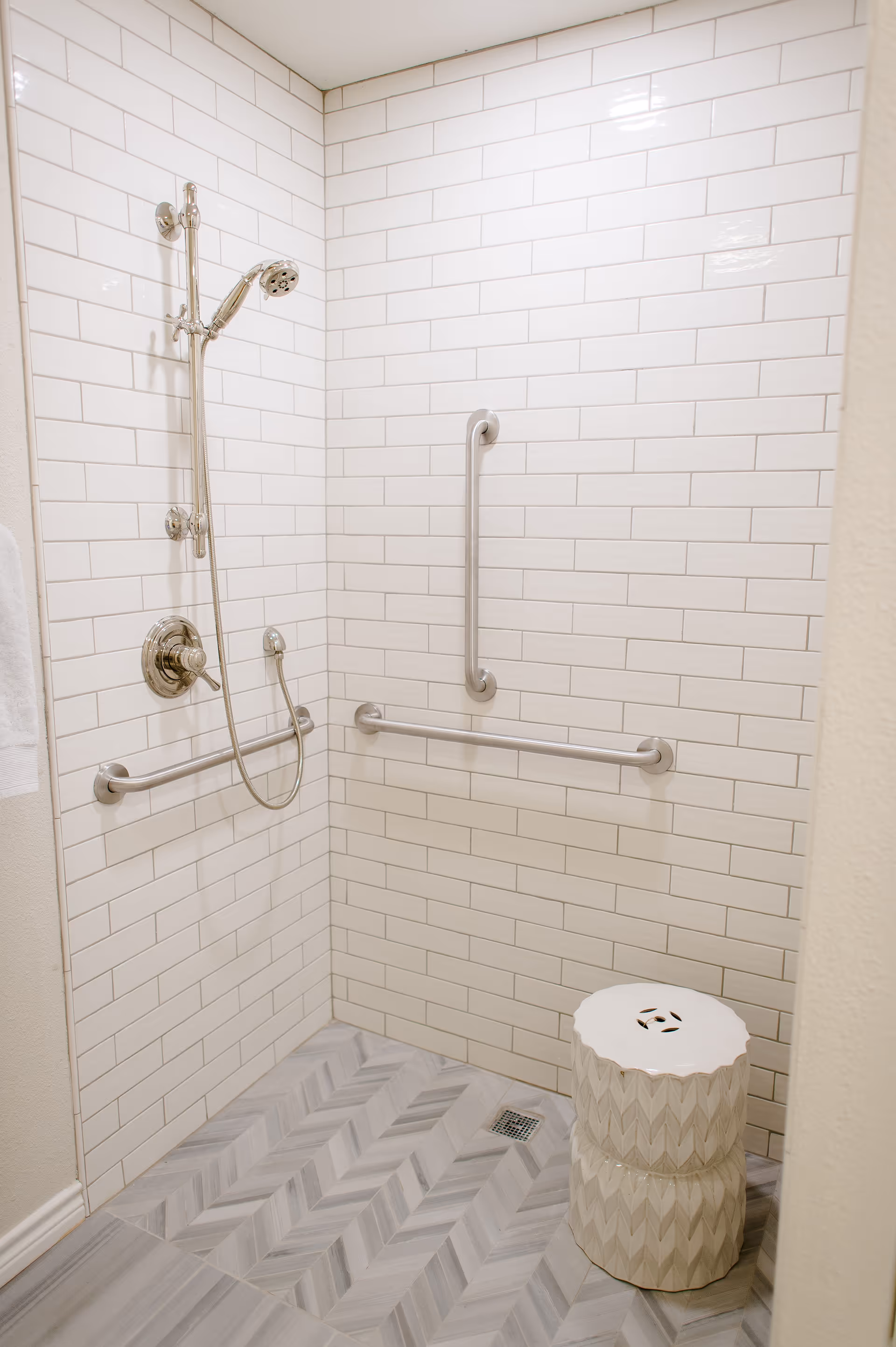 A walk-in shower with white subway tile walls and gray patterned floor tiles. The shower features a handheld showerhead, two horizontal grab bars, and a vertical grab bar for support. There is a white decorative stool inside the shower.