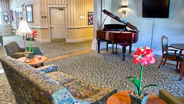 A cozy living room area in an assisted living facility featuring a patterned sofa and armchair, a round wooden table with a potted red flower, a grand piano against the wall, a wall-mounted TV, and additional seating with chairs and a small table. The room has carpeted flooring and wallpapered walls with framed pictures and a door in the background.