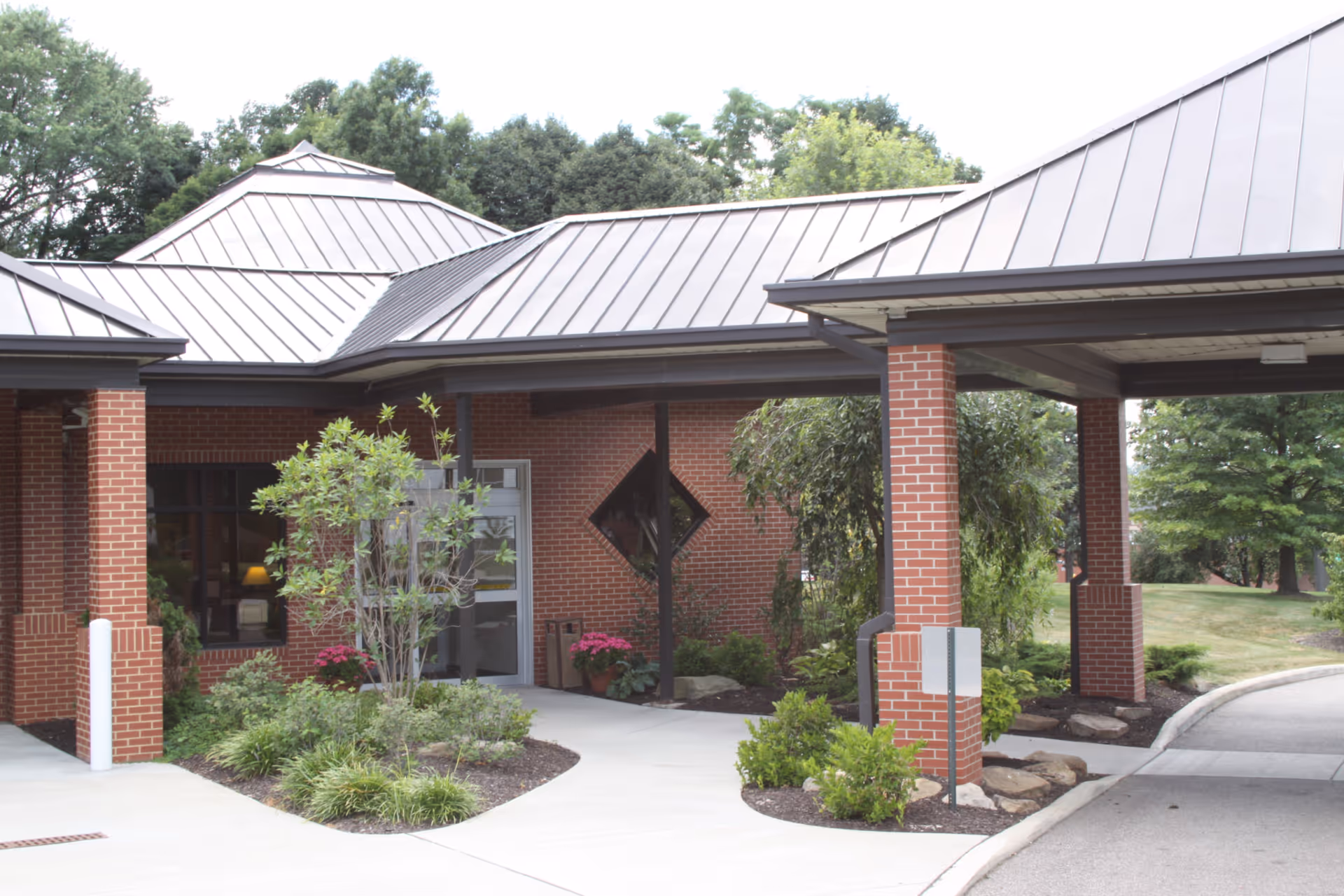 Entrance area of a brick building with a covered driveway supported by brick columns, surrounded by landscaped greenery and trees.