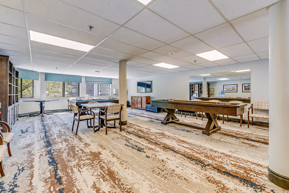 A spacious recreational room with a shuffleboard table, several chairs, and tables. The room has large windows with blue blinds, a patterned carpet, a wall-mounted TV, and a large mirror on one wall reflecting part of the room.