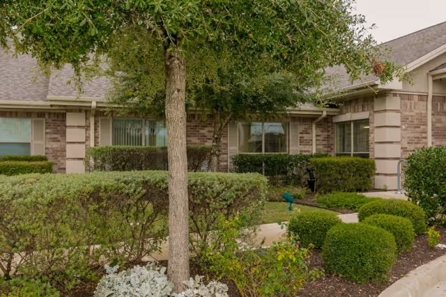 Exterior view of a brick building with multiple windows, surrounded by well-maintained bushes, shrubs, and a tree in the foreground. The scene shows a landscaped garden area with a concrete walkway.