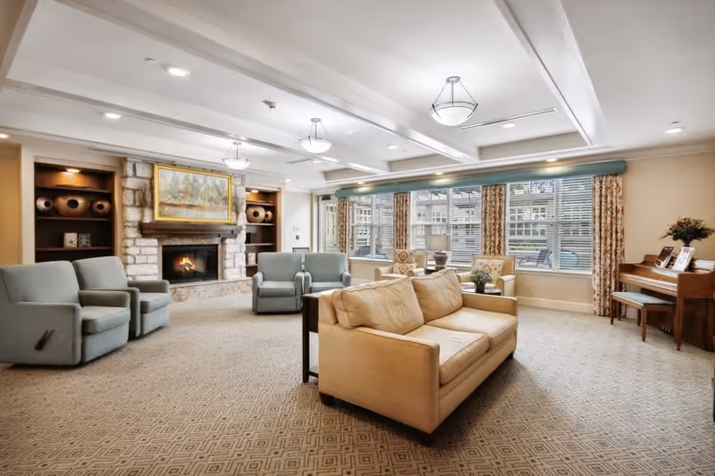 A spacious and well-lit living room with a beige sofa, four blue armchairs, a stone fireplace with a painting above it, built-in shelves with decorative items, large windows with patterned curtains, and a wooden piano with a bench on the right side.