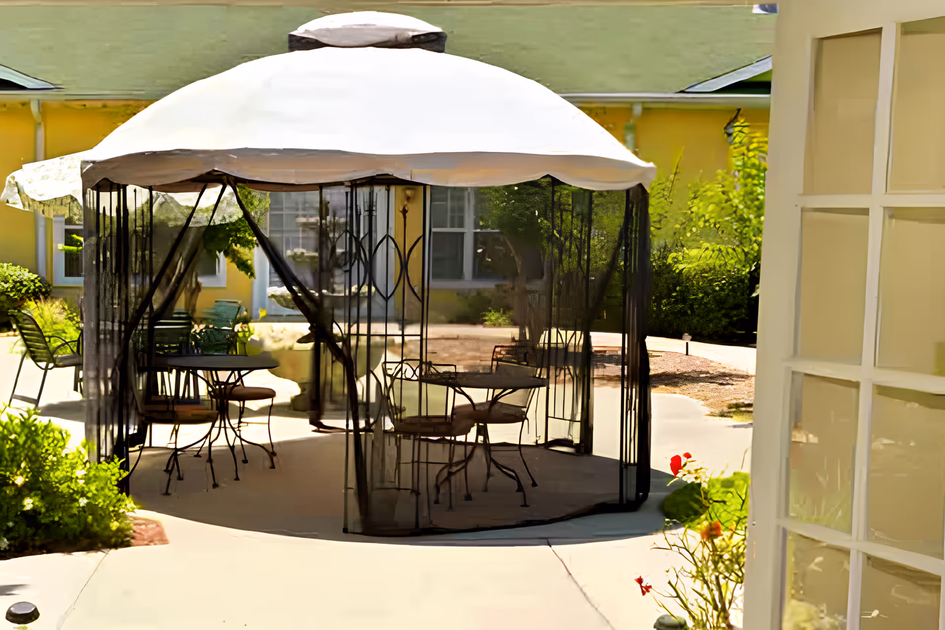 Outdoor patio area with a white canopy tent featuring black mesh sides. Inside the tent are metal tables and chairs with cushions. Surrounding the patio are green bushes and a yellow building in the background.