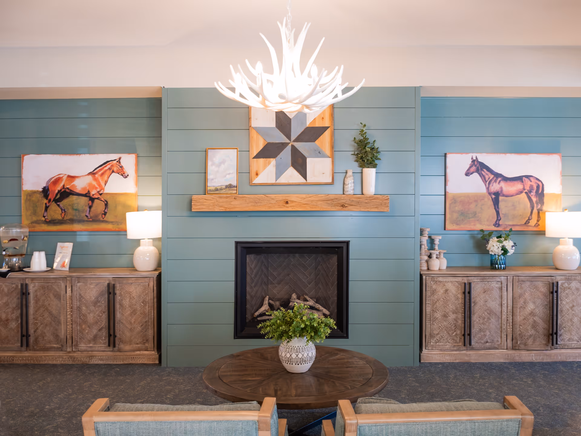 A cozy living room area with a central fireplace surrounded by teal shiplap walls. Above the fireplace is a wooden mantel with decorative items and a geometric wall art piece. On either side of the fireplace are wooden cabinets topped with table lamps, horse paintings, and decorative vases. In front of the fireplace is a round wooden coffee table with a potted plant, and two chairs with light wood frames and cushions are partially visible.