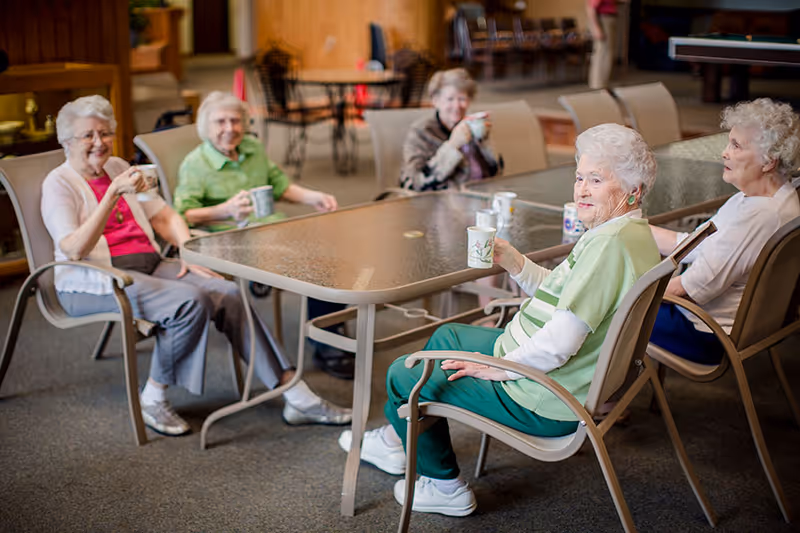 Five elderly women sitting around a large rectangular table in a common area, each holding a cup and smiling. The room has carpeted flooring and wooden paneling in the background, with additional chairs and tables visible.