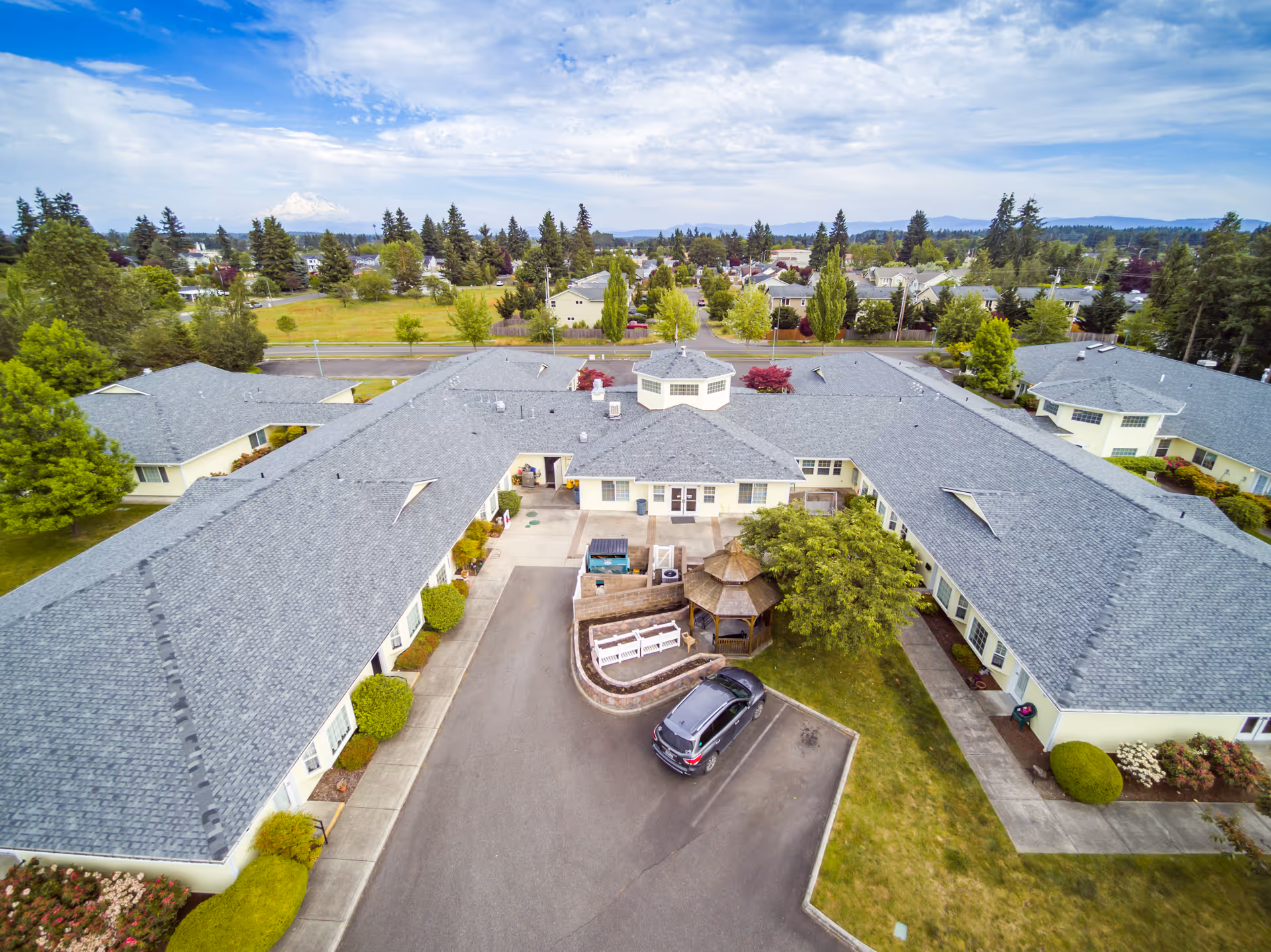 Aerial view of Easthaven Villa Assisted Living & Memory Care facility showing a large, single-story building with a gray roof arranged in a U-shape around a central courtyard. The courtyard features a gazebo, benches, and a parked car. Surrounding the building are trees, shrubs, and a residential neighborhood with more trees and houses in the background under a partly cloudy sky.