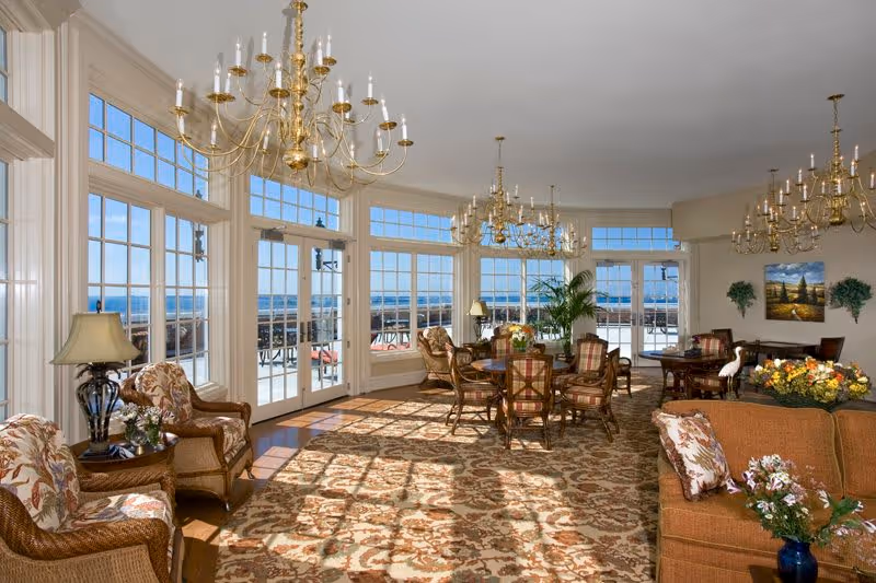 A spacious and elegant living room with large windows offering a panoramic view of the ocean. The room features multiple chandeliers, wicker armchairs with floral cushions, round wooden tables with chairs, a patterned carpet, and decorative plants and flowers. The bright natural light fills the room, creating a warm and inviting atmosphere.