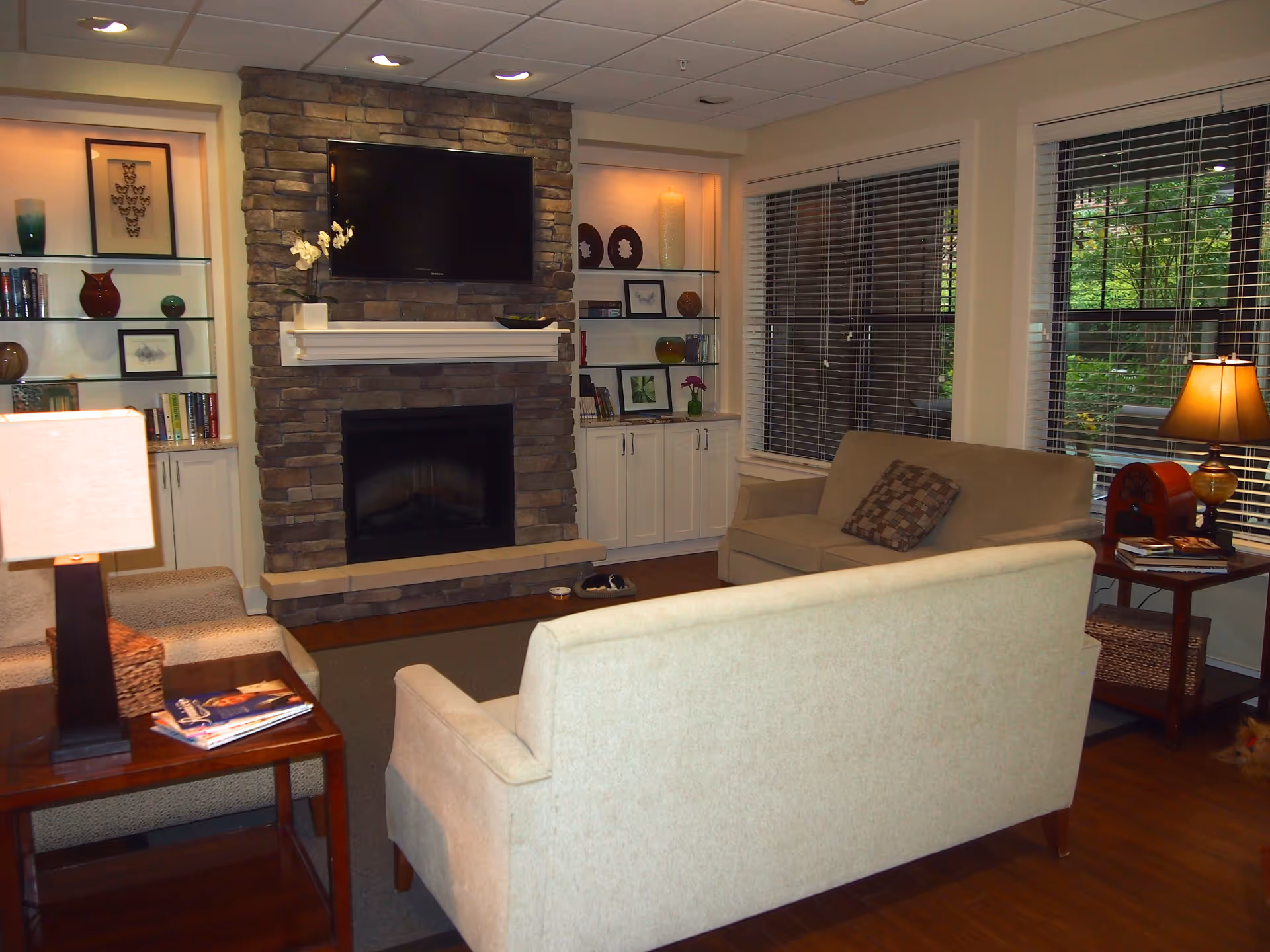A cozy living room with a stone fireplace and a mounted flat-screen TV above it. The room features two beige sofas facing the fireplace, a wooden side table with a lamp and magazines, built-in white shelves with decorative items and books, and large windows with blinds showing greenery outside.