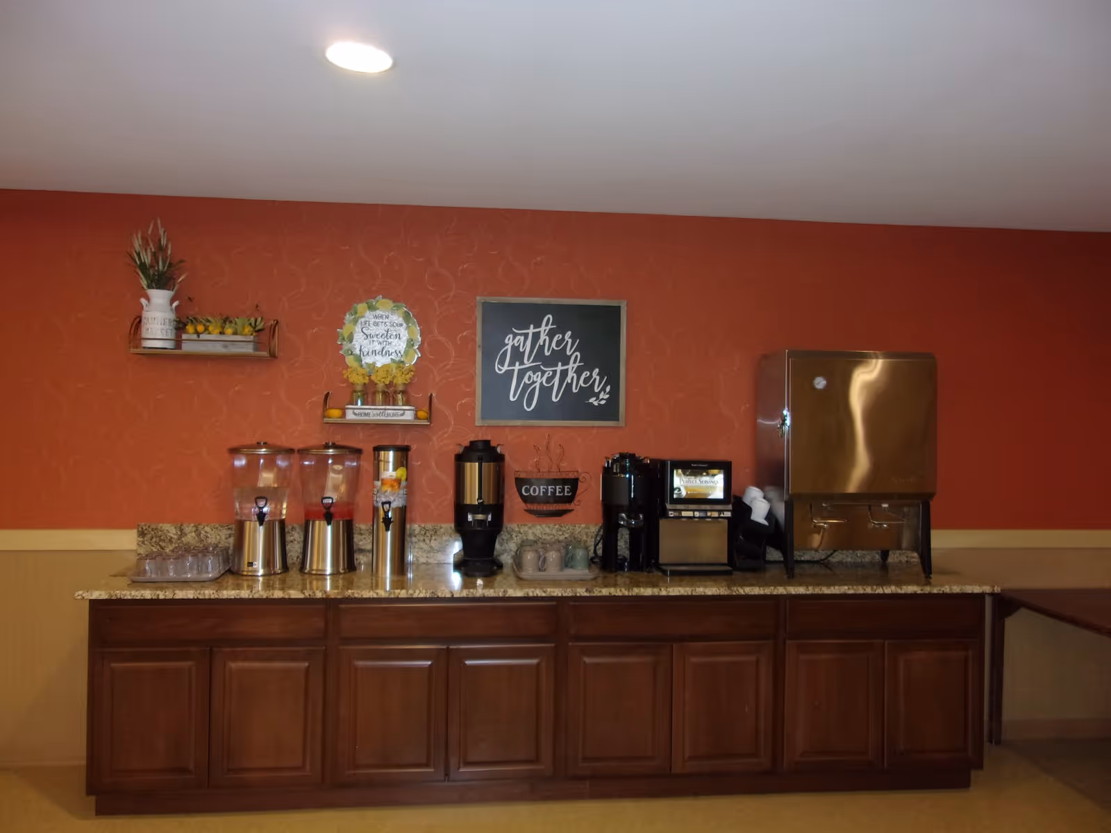 A beverage station with multiple drink dispensers, a coffee maker, and a hot water dispenser on a granite countertop with wooden cabinets below. The wall behind is painted orange with decorative signs including one that says 'gather together'.