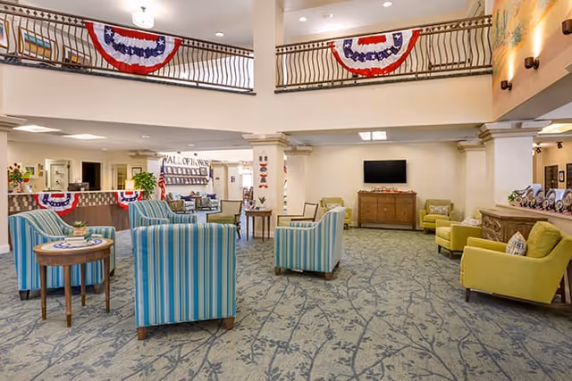 Spacious senior living facility common area with blue and green upholstered armchairs arranged around small wooden tables on a patterned carpet. The room features a high ceiling with a second-floor balcony decorated with red, white, and blue bunting. There is a television mounted on the far wall above a wooden cabinet, and various plants and decorations are visible throughout the space.
