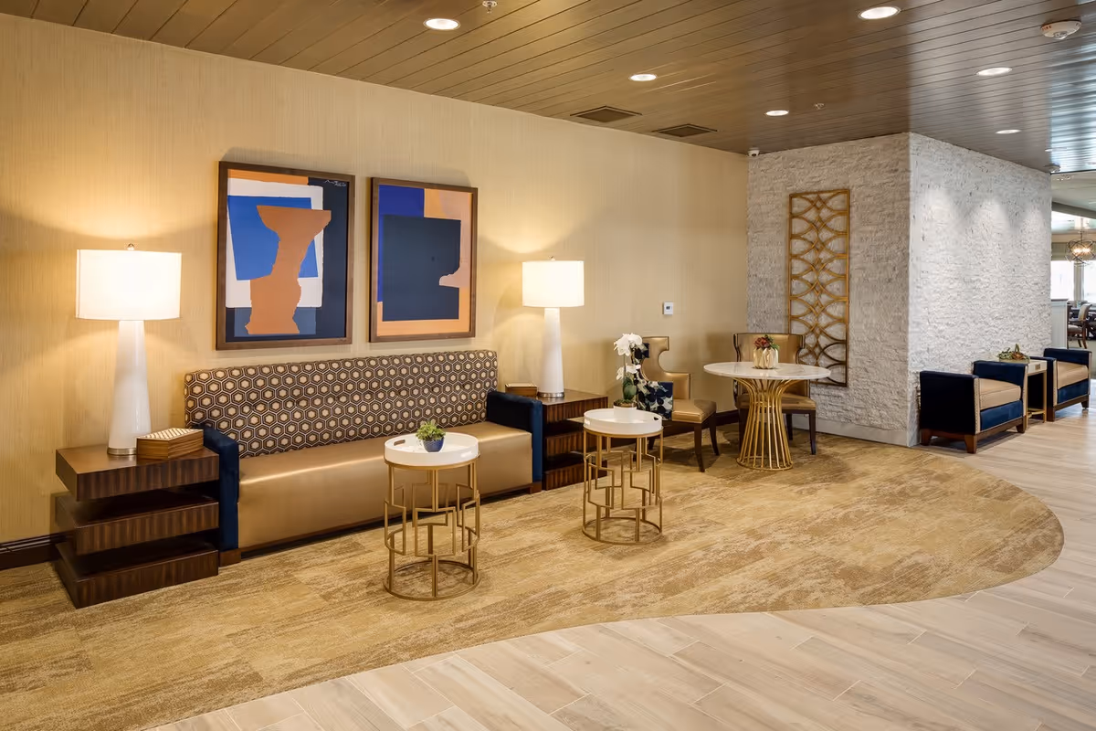A modern and stylish living room area with a patterned sofa, two white table lamps on wooden side tables, two small round white and gold tables with plants, a round white and gold table with two chairs, and additional seating along a white textured wall. The room features beige walls, a wooden ceiling with recessed lighting, and light wood flooring with a beige carpet.
