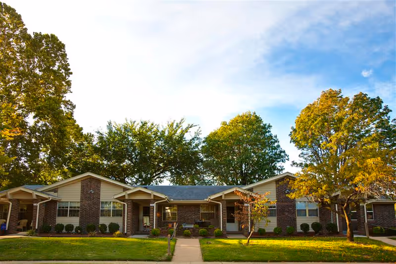 Front exterior of a single-story brick retirement community building with lawns, trees, and a central walkway.