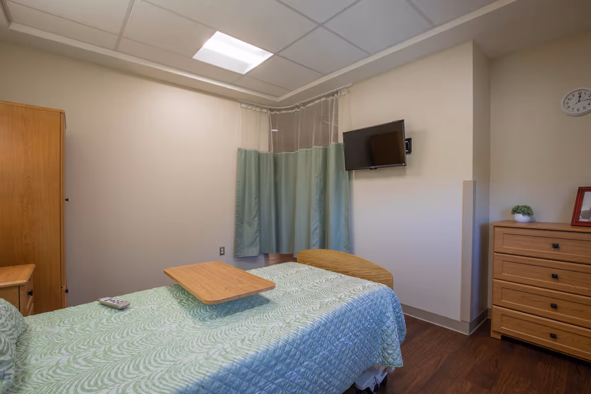 A simple nursing facility bedroom with a single bed covered in a green patterned quilt, a wooden overbed table, a wall-mounted flat screen TV, a wooden dresser with a small plant and framed picture on top, a wooden wardrobe, and a clock on the wall. The room has light-colored walls and a ceiling light panel.