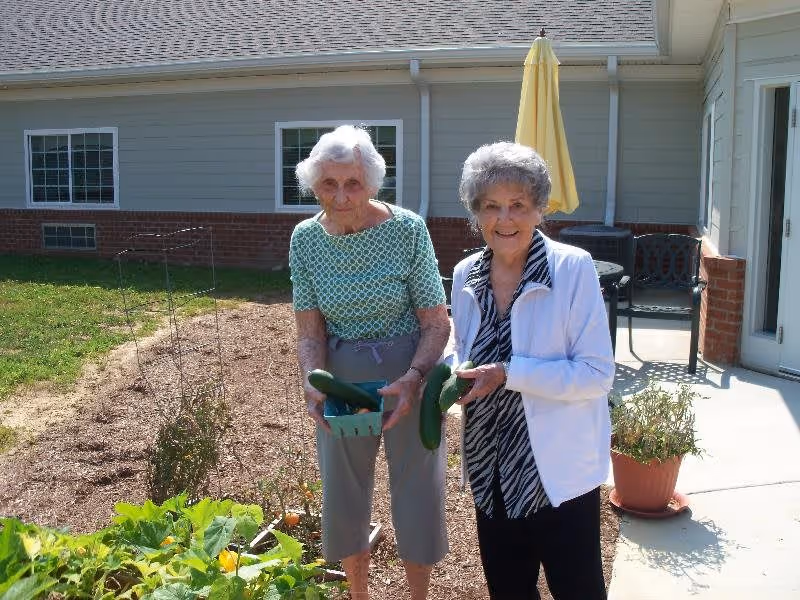 Two elderly women standing outside in a garden area next to a building. One woman is holding a basket with cucumbers, and the other is holding a cucumber. There are plants growing in the garden bed beside them, and a patio area with a table, chairs, and a yellow umbrella is visible in the background.