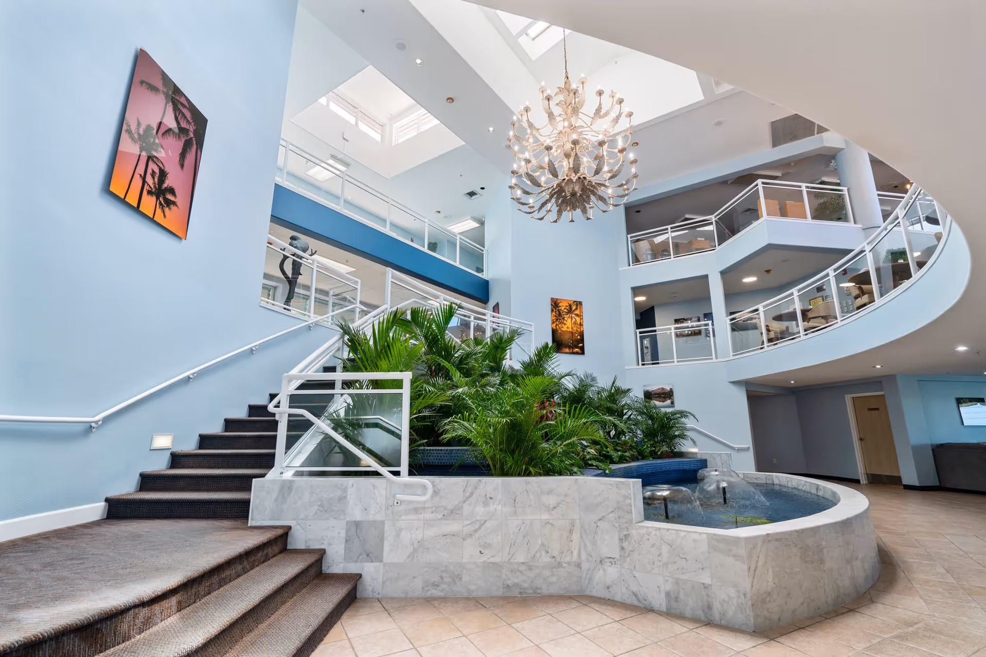 Interior view of a spacious, modern senior living facility lobby with light blue walls, a large chandelier, indoor plants, a water fountain, and multiple staircases leading to upper floors with glass railings. There are framed pictures on the walls and natural light coming from skylights above.