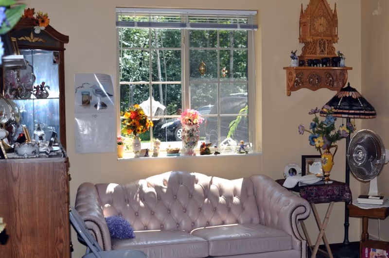 A cozy living room area with a tufted beige leather sofa placed against a wall under a window. The window sill is decorated with various flower vases and small decorative items. To the left, there is a wooden cabinet with glass shelves displaying more decorative pieces. On the right side, there is a small table with a floral lamp, a fan, and a few framed pictures. The room is warmly lit by natural sunlight coming through the window.