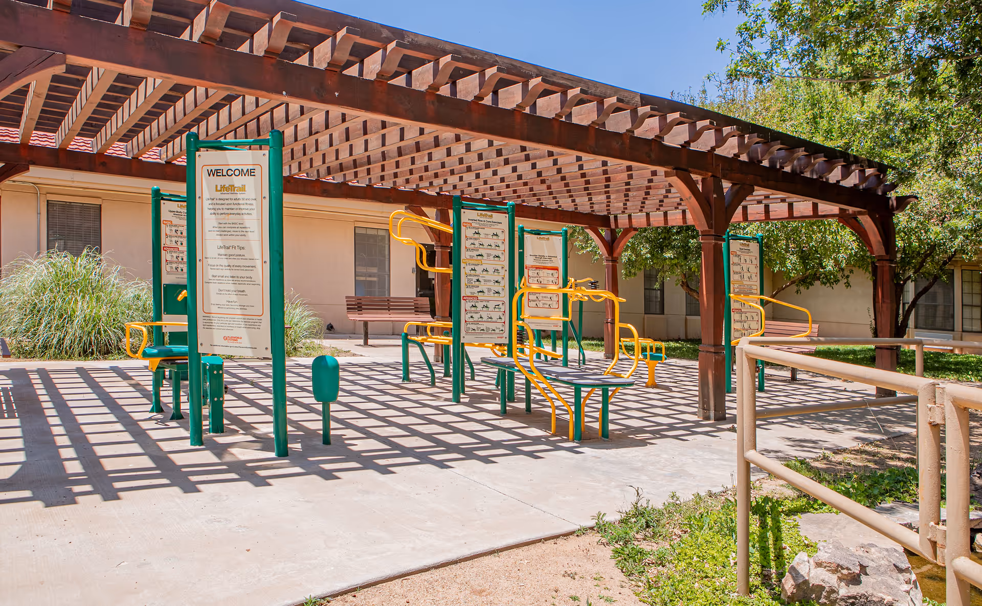 Outdoor exercise area at a senior living facility with green and yellow exercise equipment under a wooden pergola, surrounded by trees and a building in the background.