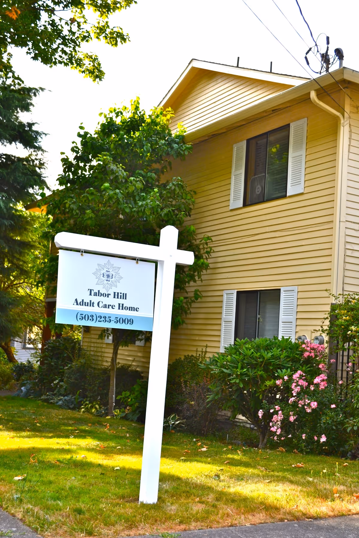 Exterior view of a yellow two-story house with white window shutters surrounded by green trees and bushes. A white signpost in the front yard reads 'Tabor Hill Adult Care Home (503) 235-5009'.