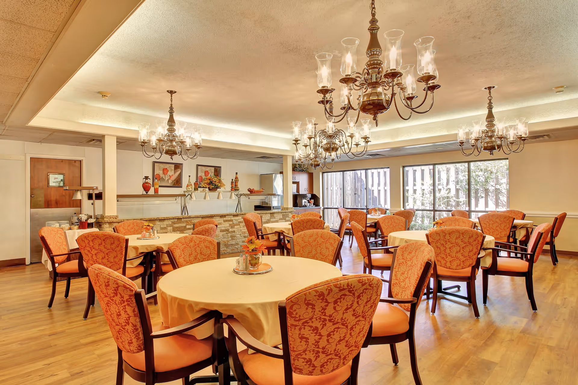 A dining room with round tables covered in beige tablecloths, surrounded by orange patterned chairs. The room features wooden flooring, large windows letting in natural light, and multiple ornate chandeliers hanging from the ceiling. There is a serving counter with decorative vases and framed artwork on the wall behind it.