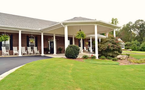 Exterior view of a single-story brick building with a covered porch supported by white columns. The porch has several white rocking chairs and hanging plants. The surrounding area features a well-maintained green lawn and some shrubs and trees in the background.