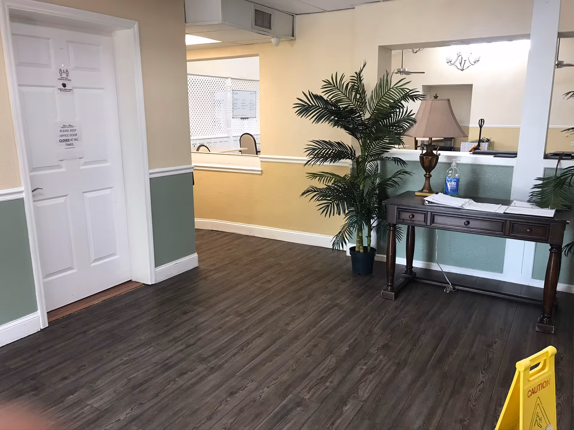 Interior view of a hallway in an assisted living facility with wood flooring, a white door with signs, a wooden table with a lamp and hand sanitizer, a potted plant, and a caution wet floor sign.