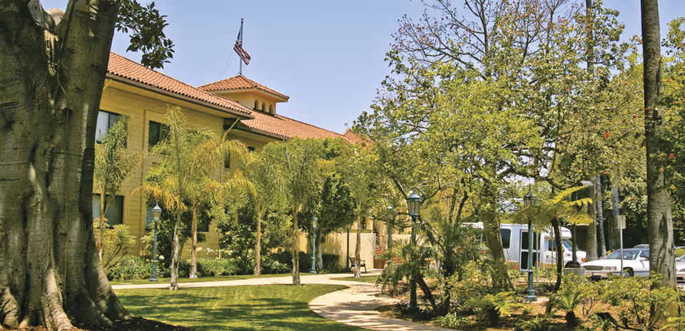 Exterior view of Hollenbeck Palms facility showing a yellow building with a red tile roof, surrounded by trees, greenery, and a curved walkway. An American flag is visible on top of the building, and several parked vehicles can be seen in the background.