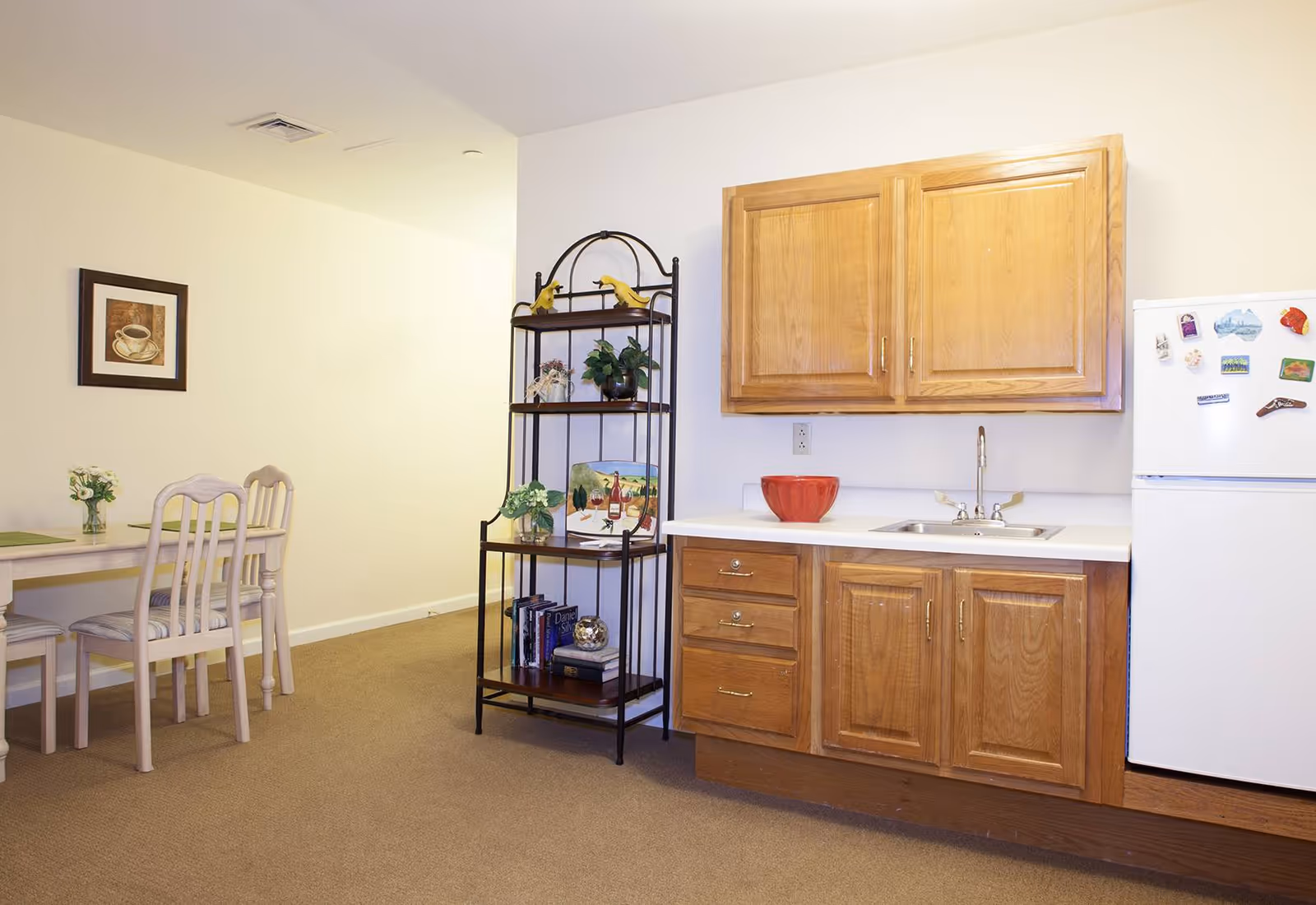 Interior view of a senior living facility kitchen area with wooden cabinets, a white countertop with a sink, a white refrigerator with magnets, and a black metal shelving unit holding decorative items and books. To the left, there is a dining table with chairs and a framed picture of a coffee cup on the wall.