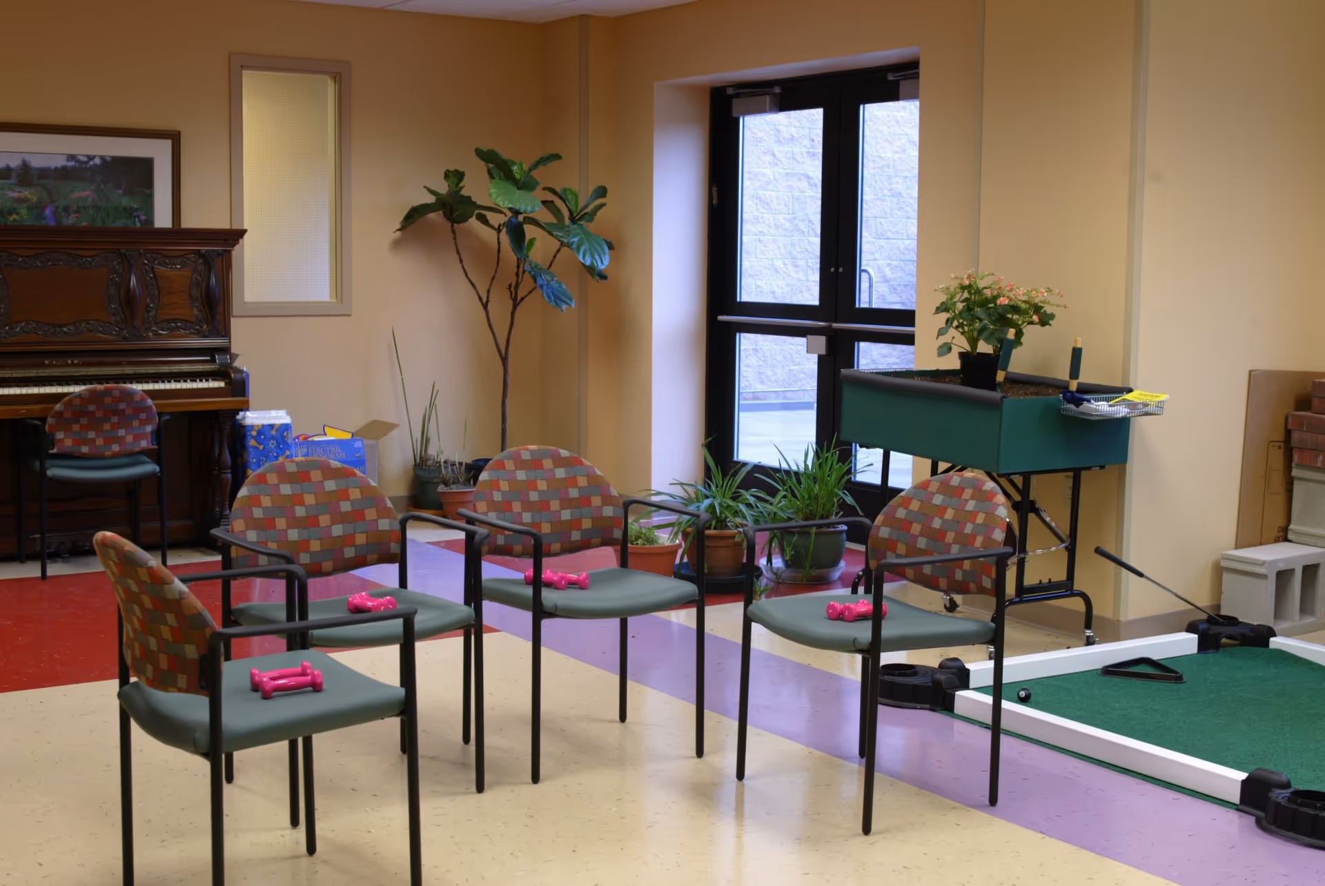A common area in an assisted living facility with four patterned chairs arranged in a circle, each holding a pair of pink dumbbells. There is a piano with a chair in the background, several potted plants near a glass door, and a green gardening table with plants on the right side. The floor has a colorful pattern with red, purple, and beige tiles.