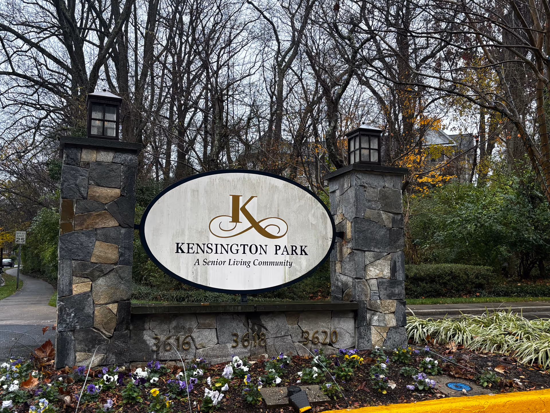 Stone entrance sign reading "Kensington Park A Senior Living Community" flanked by two lantern-topped stone pillars and landscaped flowers.