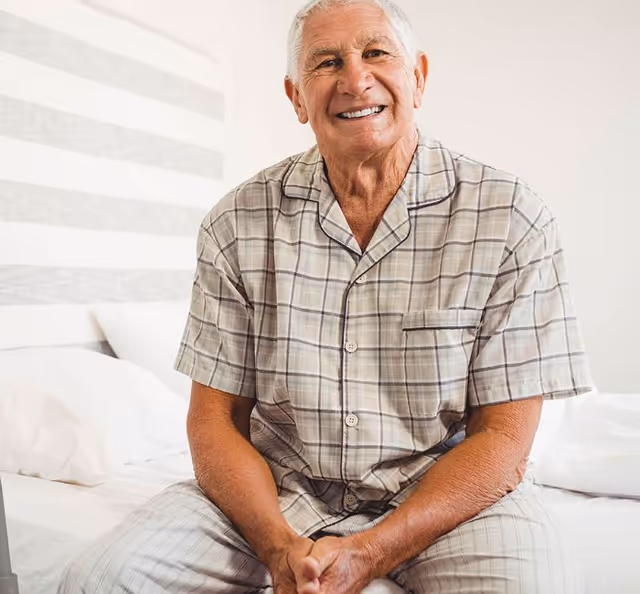 An elderly man wearing plaid pajamas sitting on a bed in a bright bedroom, smiling at the camera.
