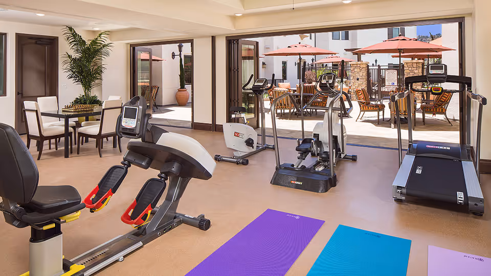 Indoor fitness room with exercise equipment including a recumbent bike, elliptical machines, and a treadmill. Three exercise mats are laid out on the floor. Large glass doors open to an outdoor patio area with tables, chairs, and umbrellas.