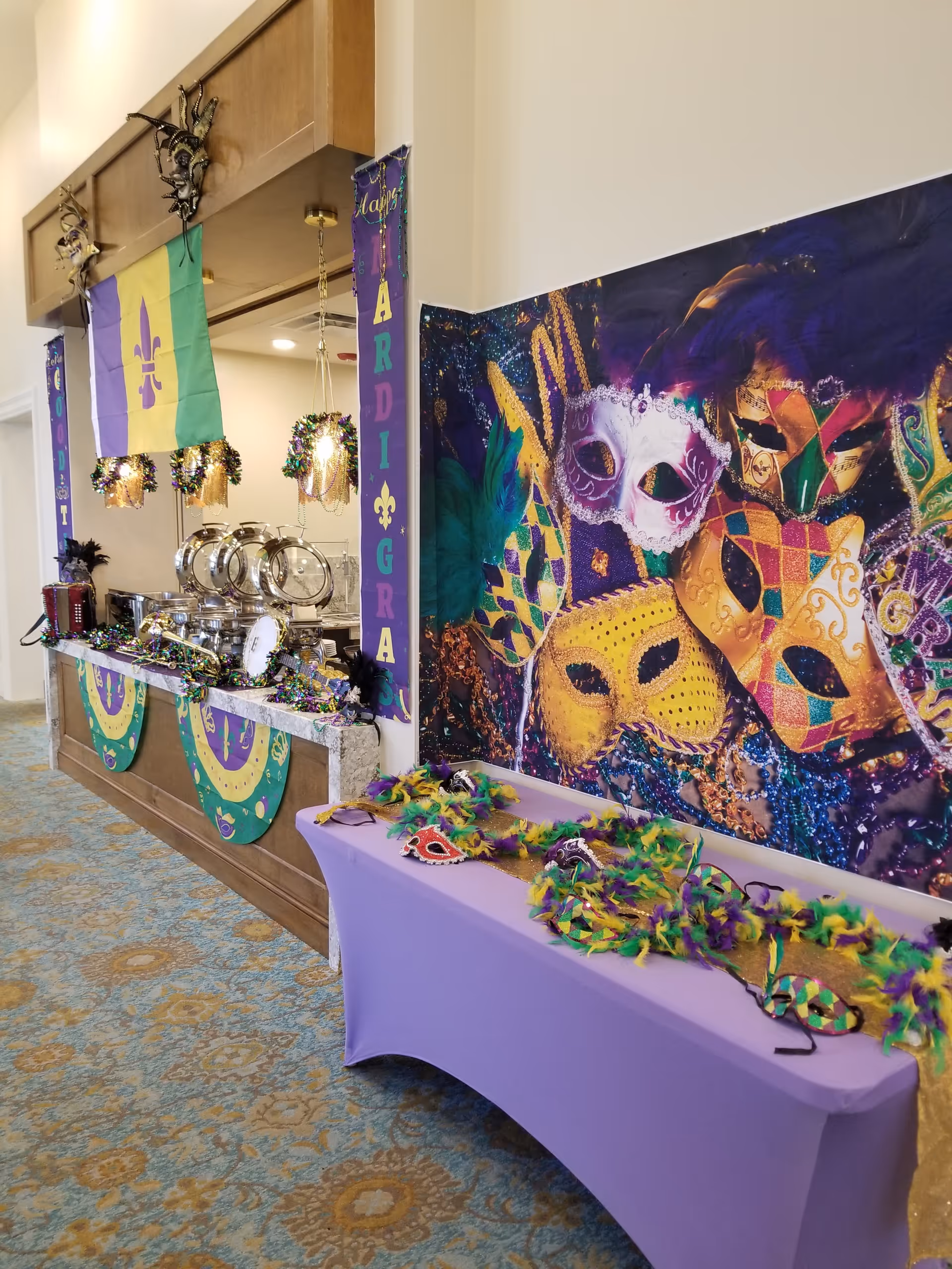 Indoor Mardi Gras-themed buffet setup with decorative masks, beads, purple tablecloth and chafing dishes along a decorated wall.