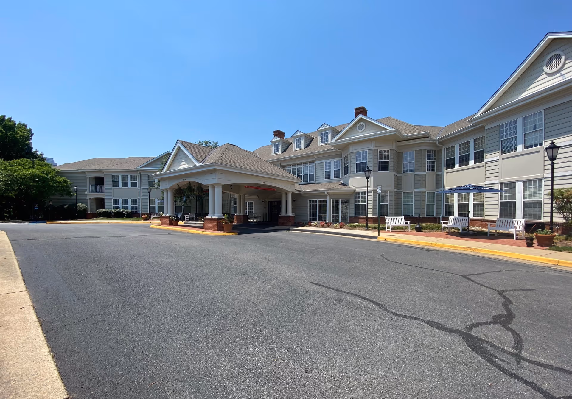Front entrance of a large multi-story senior living building with a porte-cochère, benches, and a paved driveway under a clear blue sky.