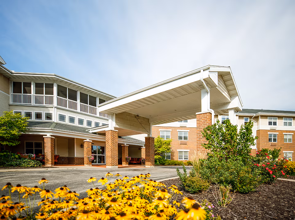 Exterior view of Hickory Park Senior Living building with a covered entrance supported by brick columns, surrounded by landscaped flower beds with yellow and red flowers under a partly cloudy sky.