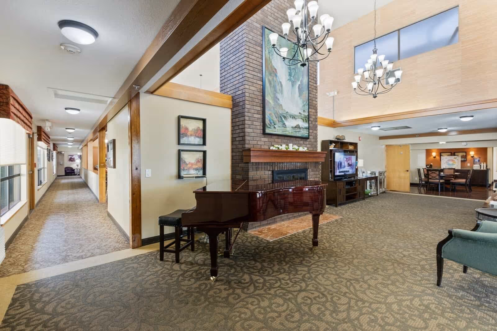 Interior view of a senior living facility common area with a grand piano in the foreground, a brick fireplace with a large painting above it, chandeliers hanging from a high ceiling, a television on a stand, and a dining area in the background. A long hallway with windows and carpeted floor extends to the left.