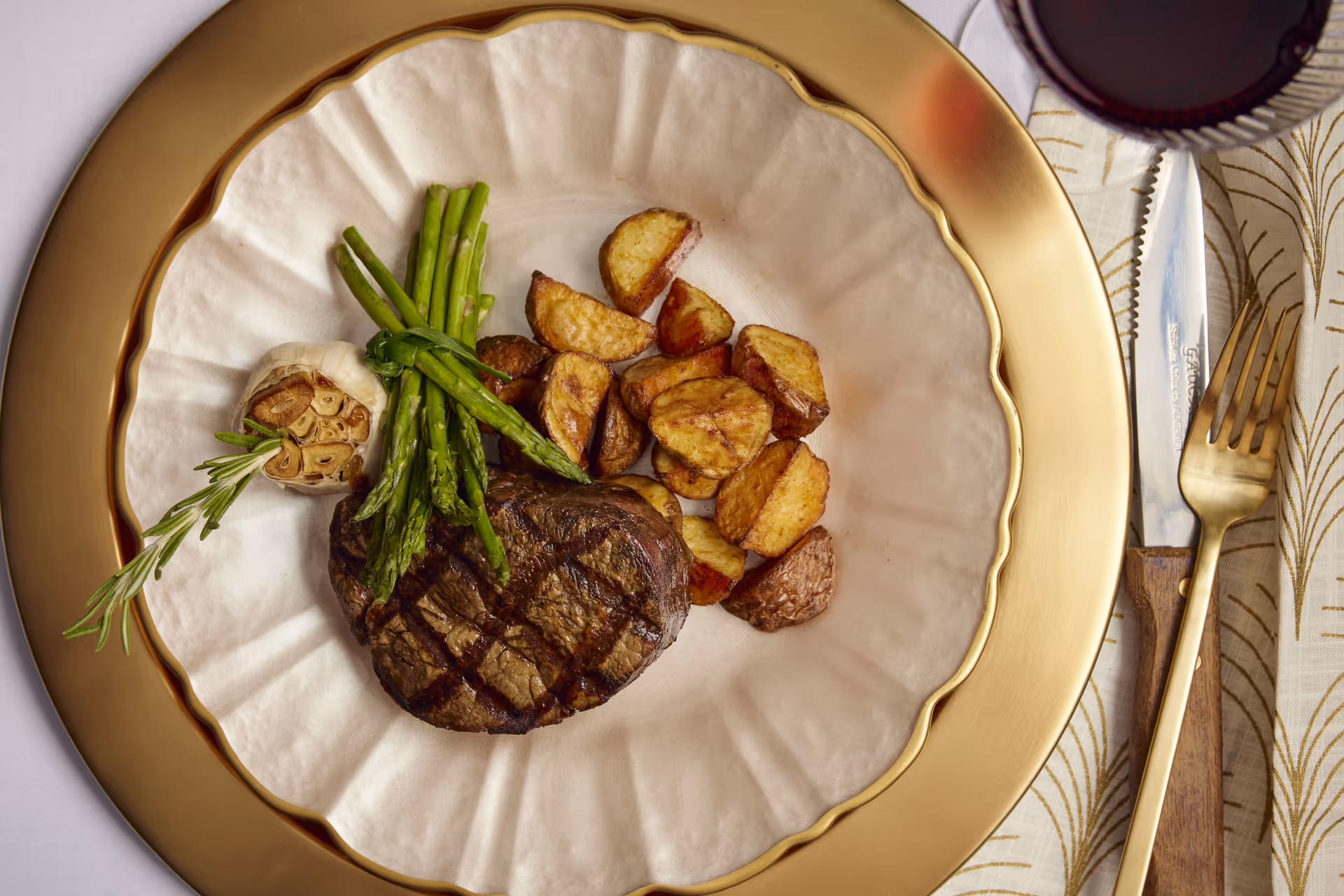 Plated steak with roasted potatoes, asparagus and a roasted garlic bulb on a gold charger with cutlery and a glass of red wine nearby.