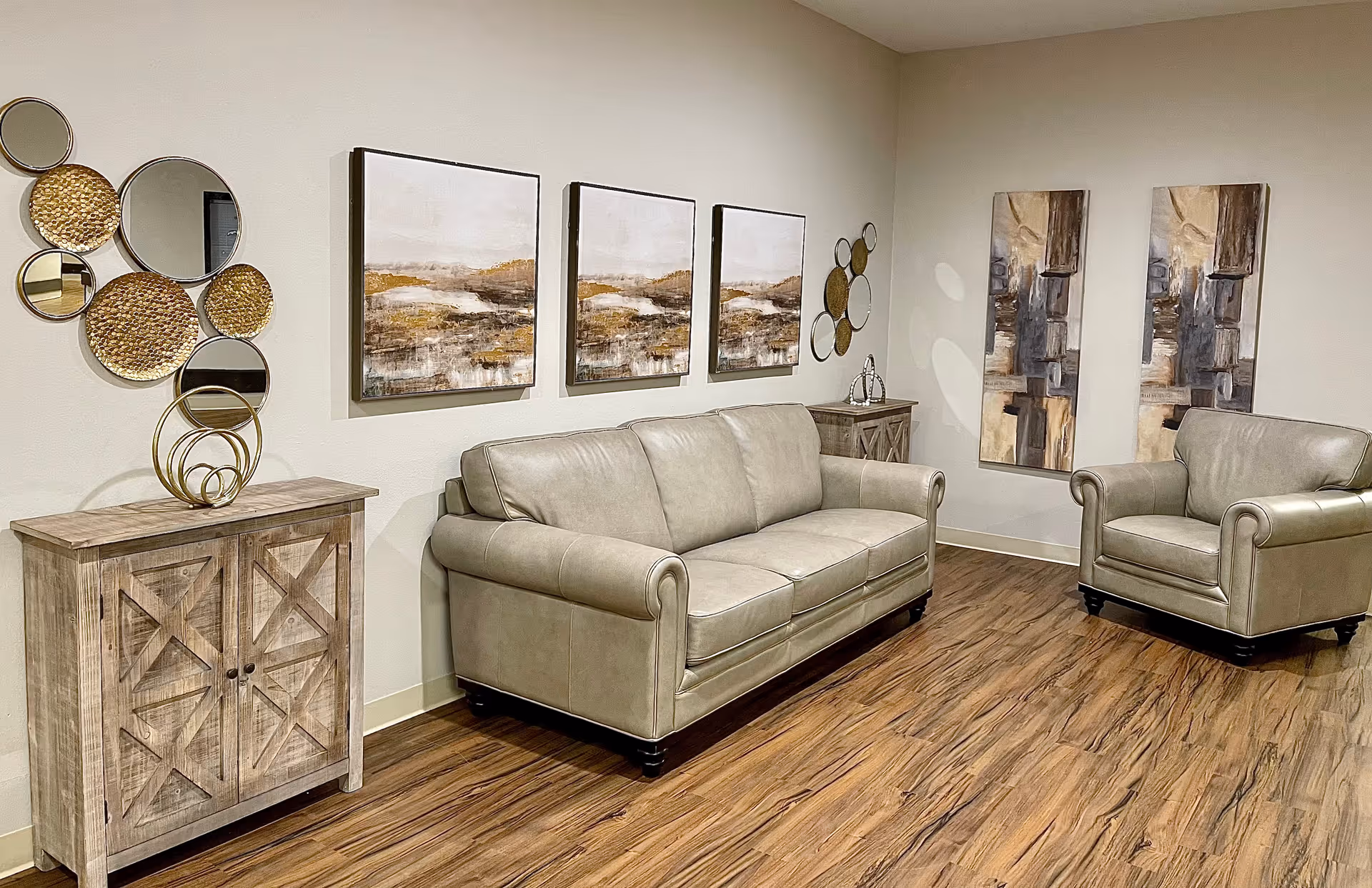 A cozy living room area with a beige leather sofa and matching armchair on a wooden floor. The walls are decorated with multiple abstract paintings and circular mirrors. There are two wooden side cabinets with decorative items on top.