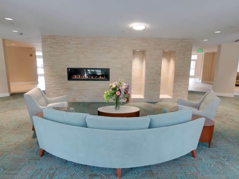 Curved seating area with a coffee table and flower arrangement facing a built-in linear fireplace in a bright lobby.