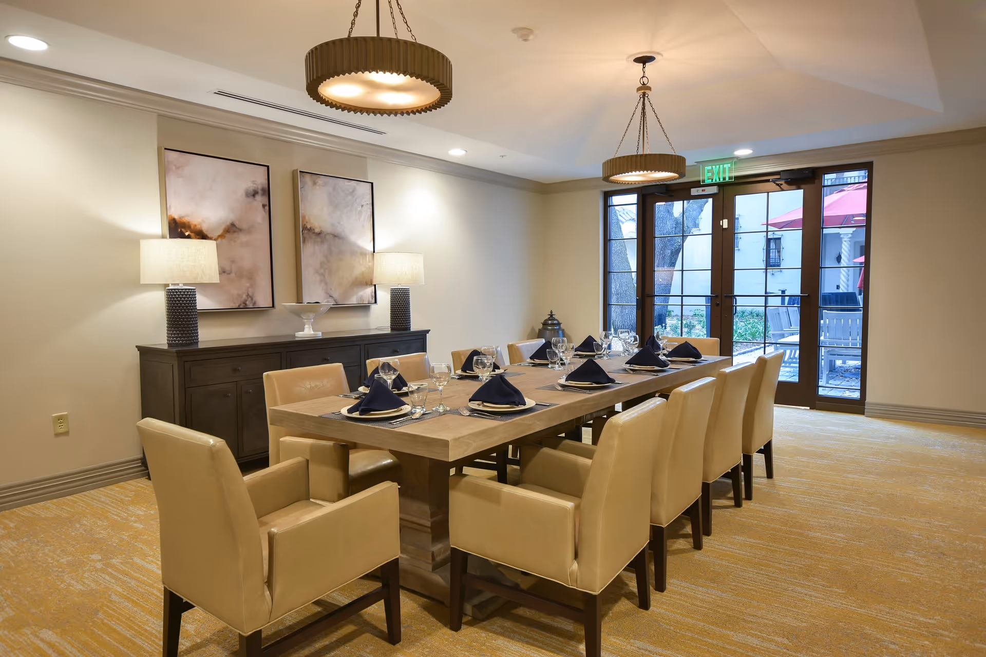 A dining room with a long wooden table set for twelve people with beige leather chairs. The table is set with plates, glasses, and black folded napkins. Two modern pendant lights hang from the ceiling. There is a dark wooden sideboard against the wall with two table lamps and two abstract paintings above it. Glass doors lead to an outdoor patio area with red umbrellas and seating.