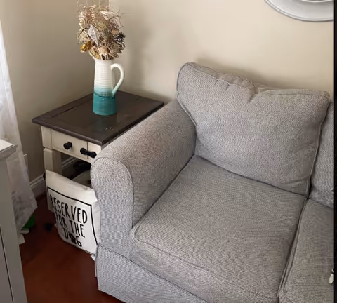 Gray upholstered armchair beside a dark wood side table with a vase of dried flowers indoors.