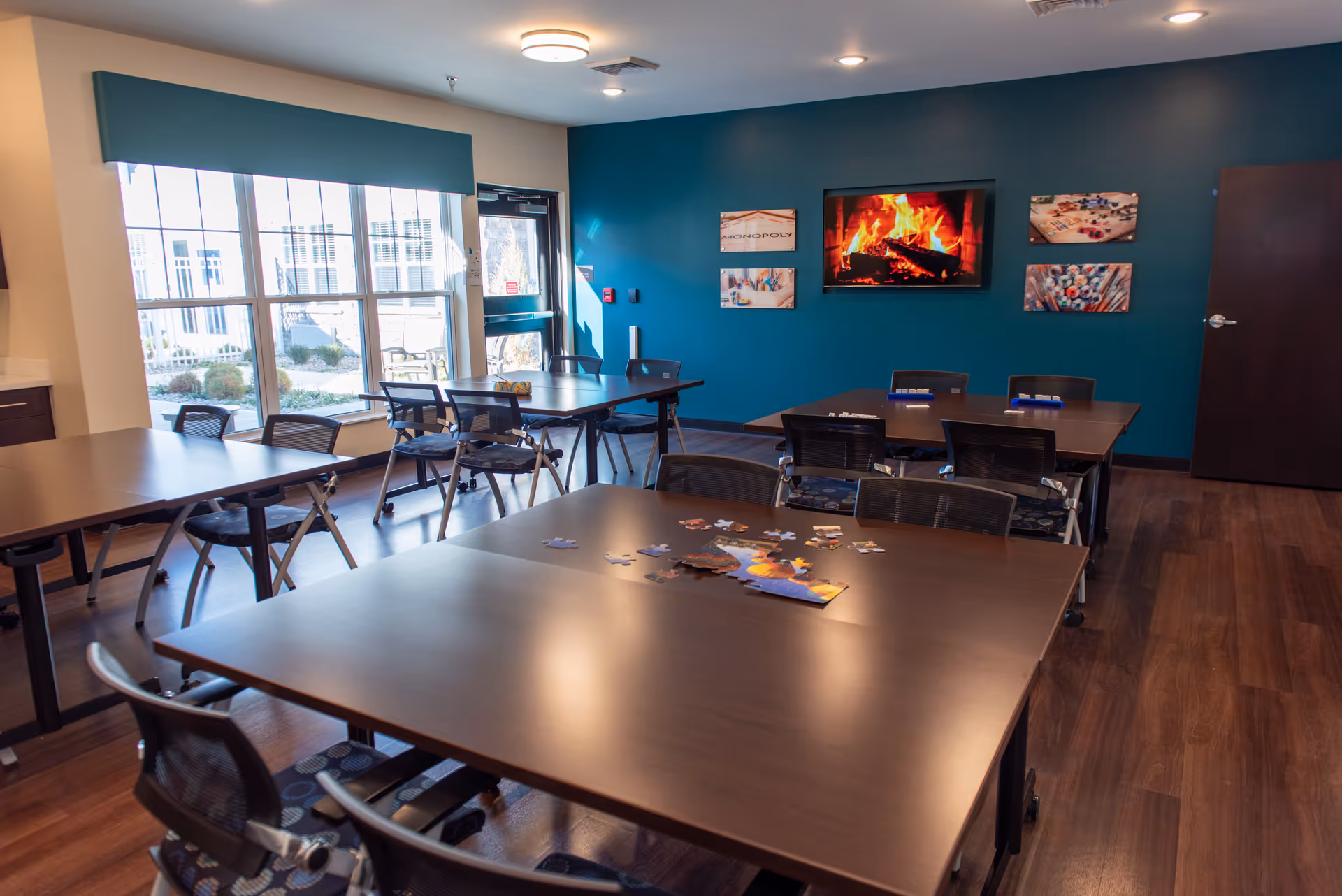 A well-lit common room with several tables and chairs arranged for group activities. A large window lets in natural light, and a door leads outside. On the teal-colored wall, a flat-screen TV displays a cozy fireplace scene, surrounded by framed pictures of board games. Puzzle pieces are scattered on the nearest table.